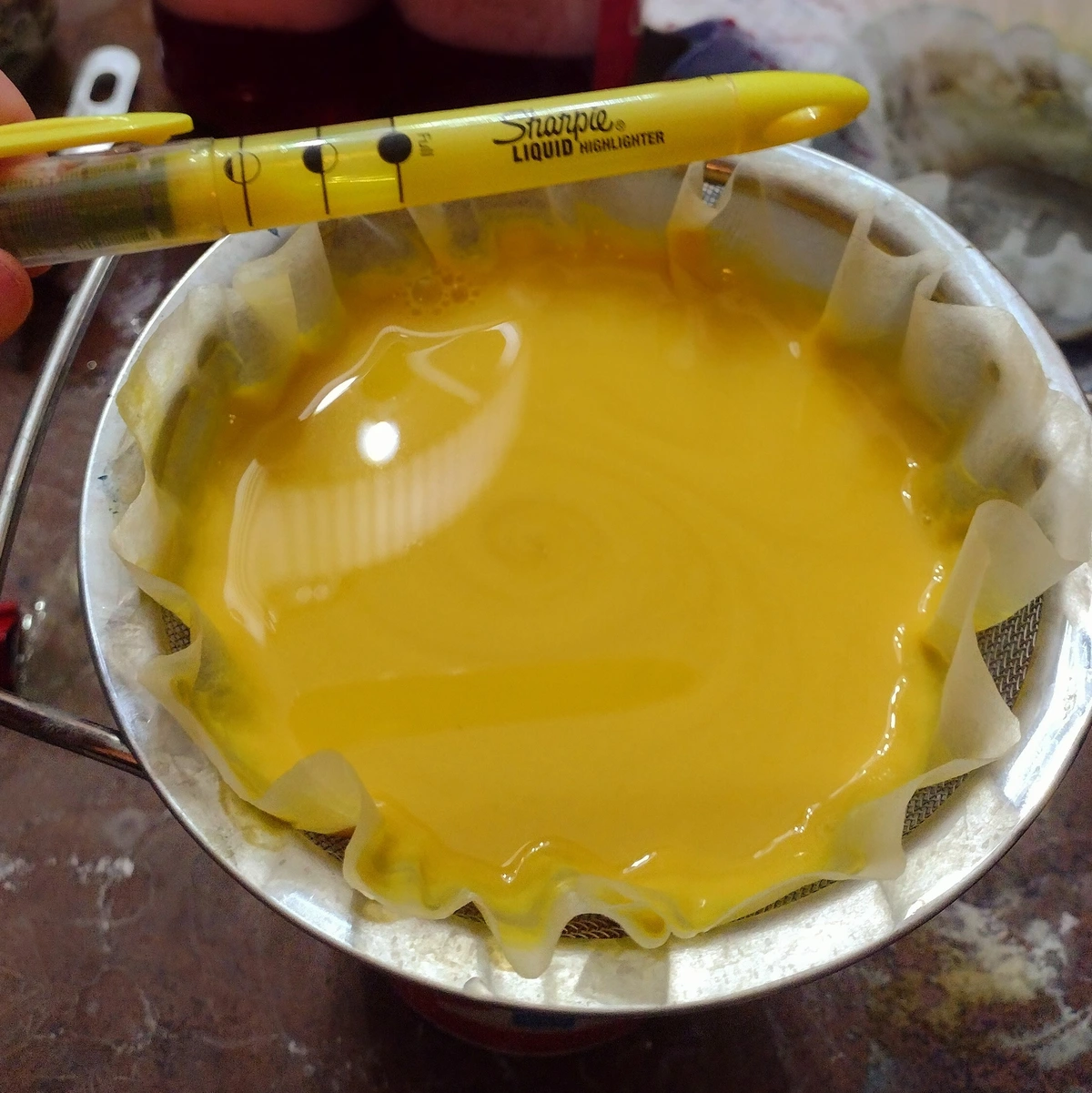 A coffee filter sitting in a strainer on top of a jar, filled with a bright yellow goop, with a yellow highlighter held over it to compare the intensity of the colour.