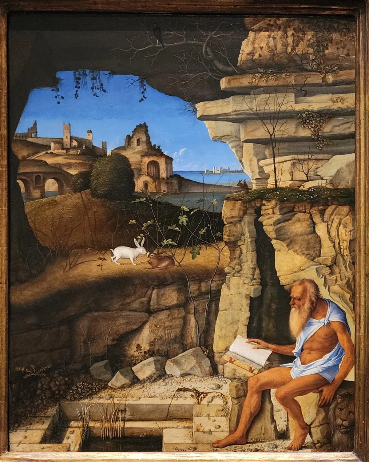 "at bottom right, an elderly Jerome clad in rags reading a book at the entrance of a cave. at his feet is a stone floor that descends in a square hole into a pool or baptistery. above the pool the rocky terrain opens into a perspective view of cities and seas in the distance. above the pool are two rabbits facing."
