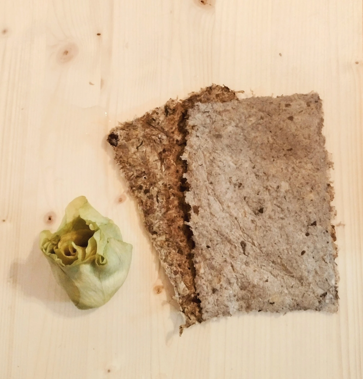 Two pieces of grayish, rough handmade paper next to a soaked yellow rose head on a wooden board.