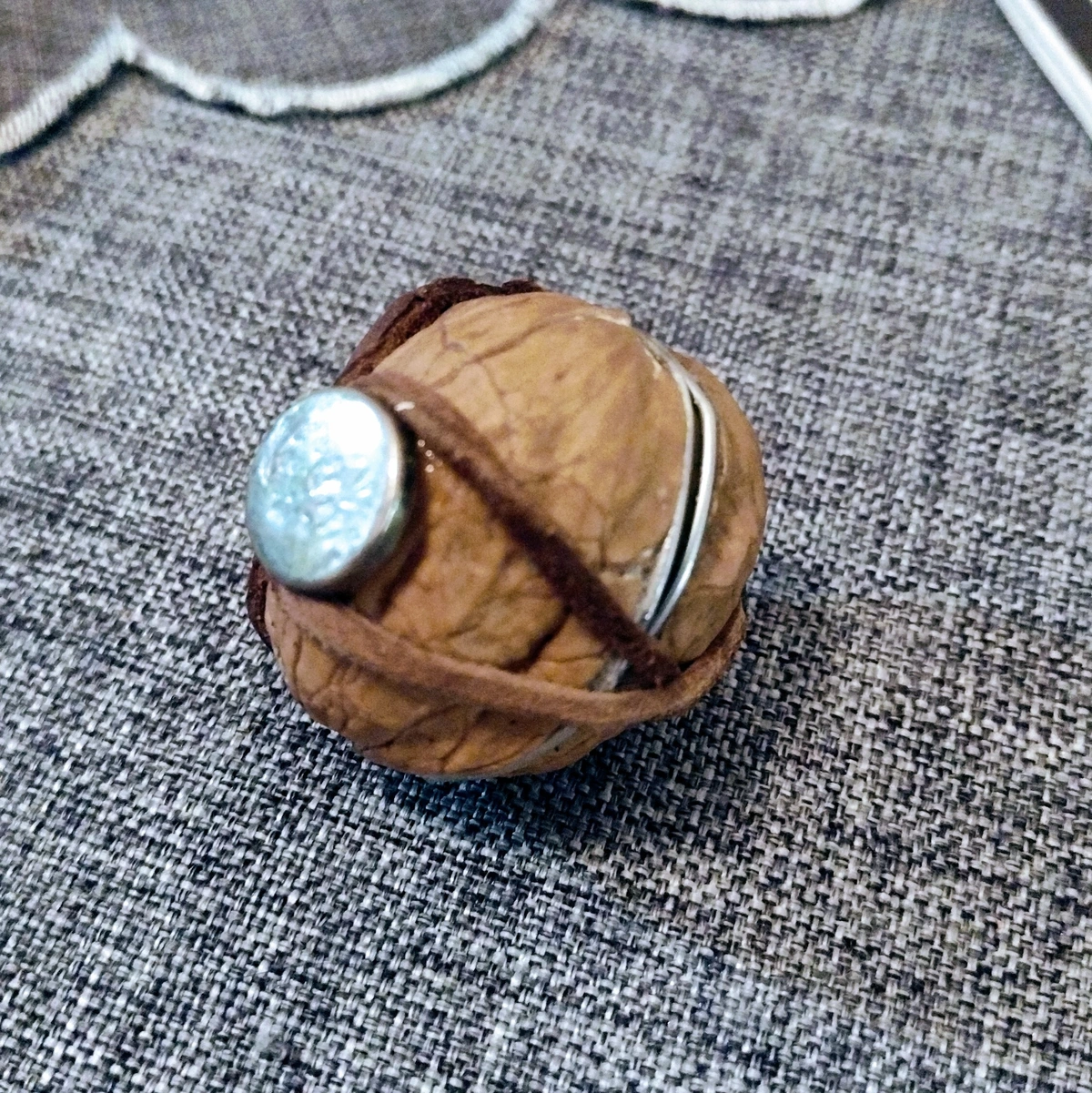 A walnut shell turned into a tiny box with a leather strap and a metal button for a closure. Sitting on a gray tablecloth.