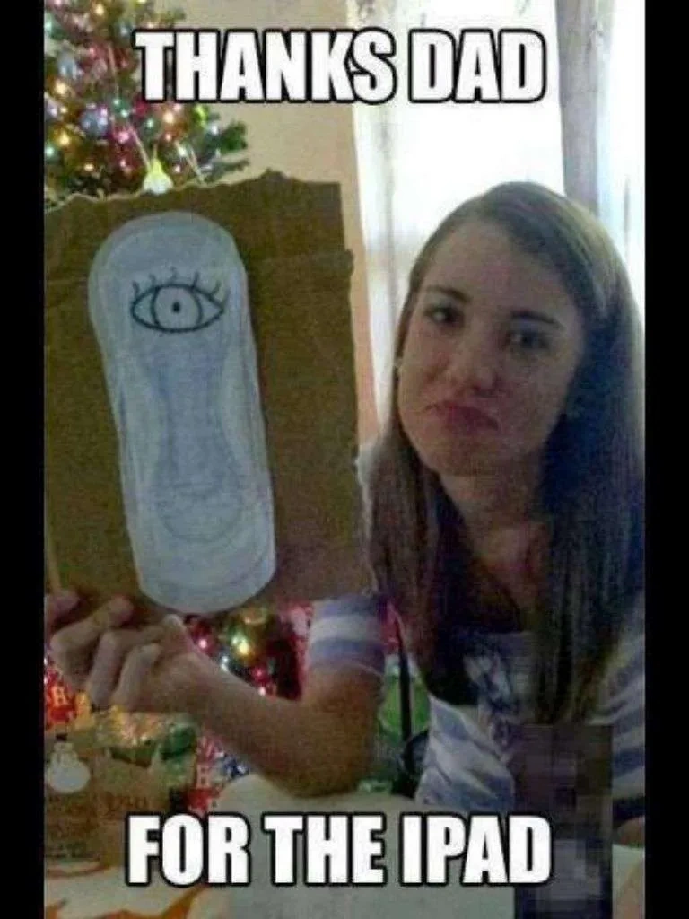 A meme of a young woman sitting in a living room in front of a decorated tree. She's holding a ripped piece of cardboard with a menstrual pad stuck on, on which is drawn a rough cartoon eye. The text reads: "Thanks dad for the ipad."