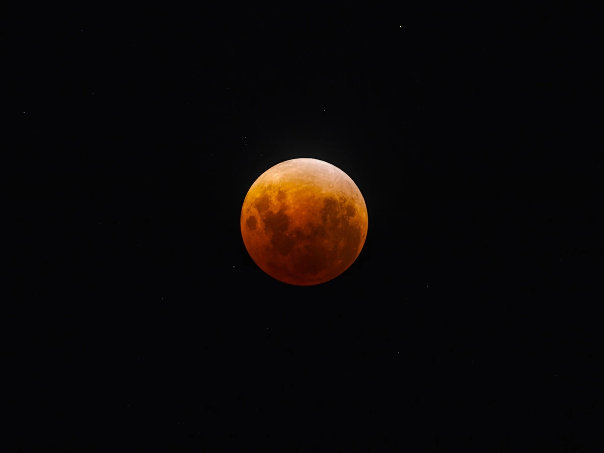 A full moon, tinted in orange and red, during the peak of the eclipse.  Stars are visible in the background of the night sky
